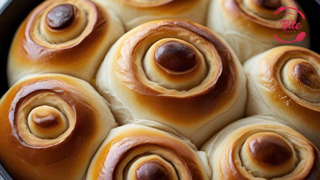 Warm Dinner Rolls Golden-brown spiral buns in a baking pan, each with a soft, creamy swirl. The image conveys warmth and appetizing appeal. Logo says "Bite Villa."