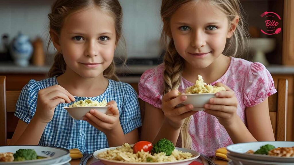 Two young girls sit at a dining table, smiling, holding small bowls of pasta. Plates with pasta, broccoli, and tomatoes are in front of them in a warm kitchen setting.